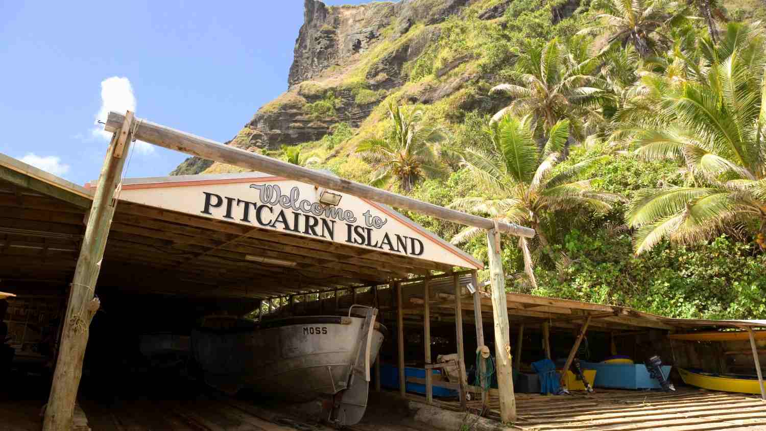 Welcome to Pitcairn Island sign above boathouse with palm trees and rugged cliffs in the background